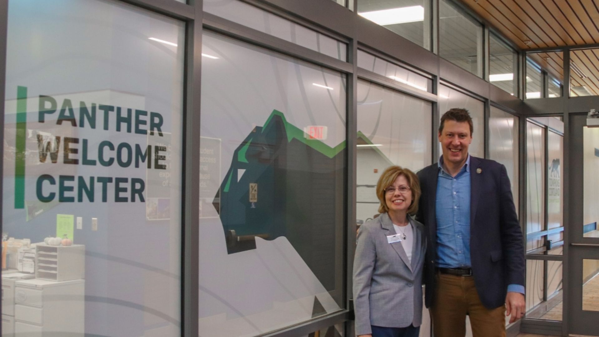Two people in formal dress stand in front of a wrapped glass wall that has Panther Welcome center on it, and a giant Panther. 