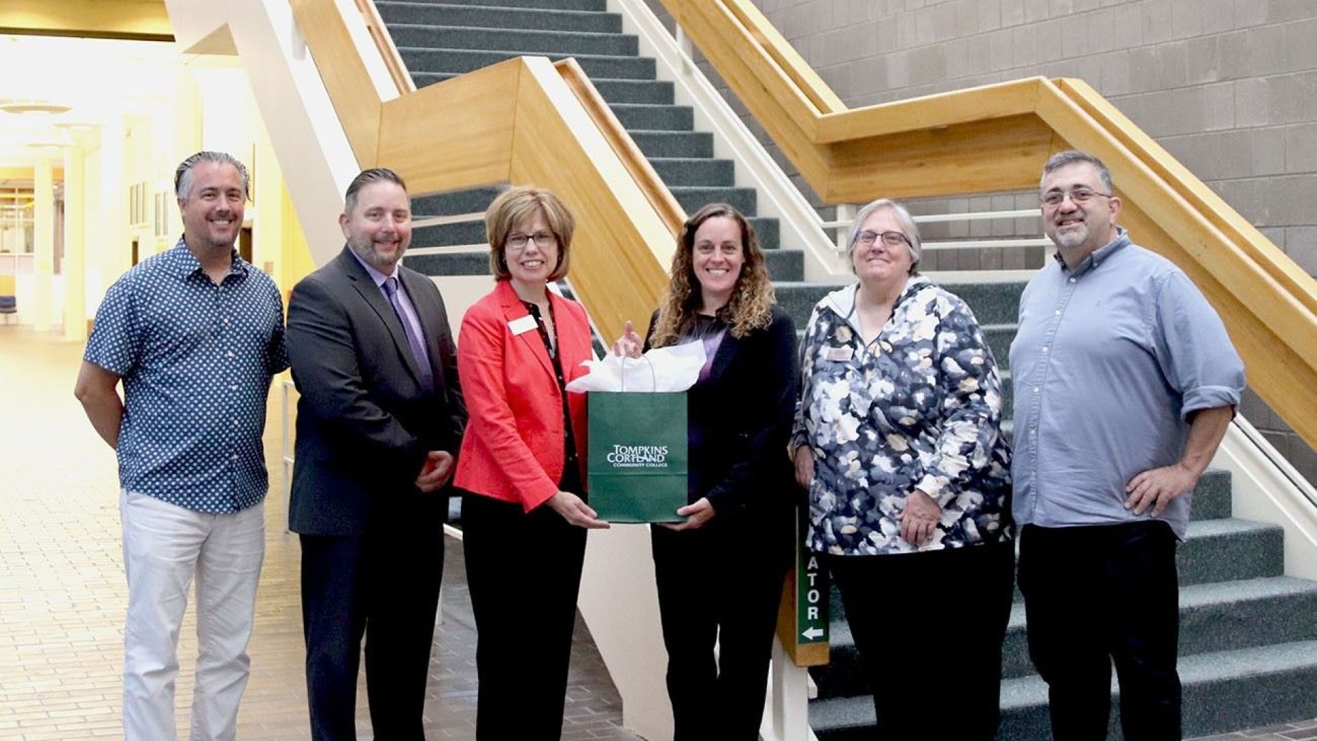 Group of six people standing in front of a staircase, with one person holding a gift bag