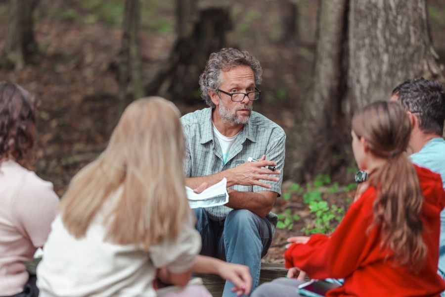  A professor sits outdoors in a forested area, engaging with a group of students seated around him, holding notes and gesturing while leading a discussion.